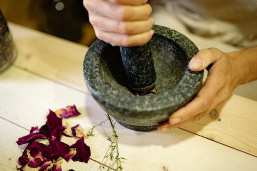 Person grinding herbs in a stone mortar and pestle on a wooden table, with dried rose petals nearby.